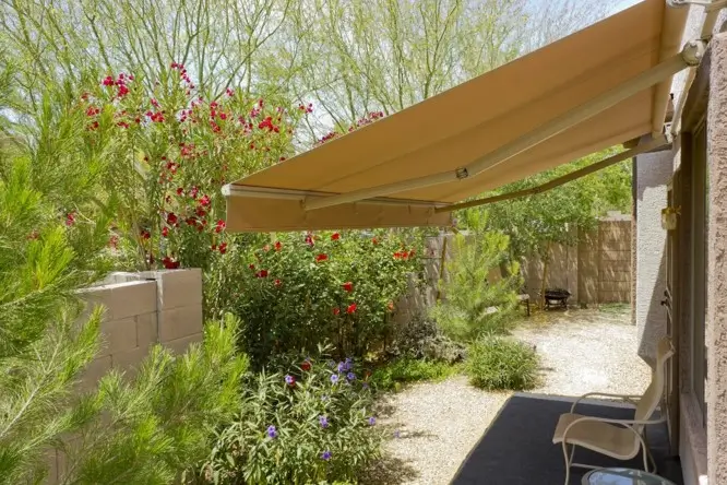 Tan folding arm awning providing shade over a small garden patio with flowering plants